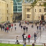 Looking down at a square enclosed on two sides by white stone castle-like buildings and in front by a green lawn with a round table-like structure; many people strolling